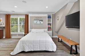 A bedroom with a large bed, a bench, a TV, a bookshelf, and a window with curtains. - Fully equipped studio in the center of town near train, beach and town green (Guilford)