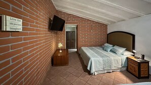 Une chambre avec un mur en briques, un téléviseur fixé au mur, un lit avec une tête de lit, une table de chevet et une commode.