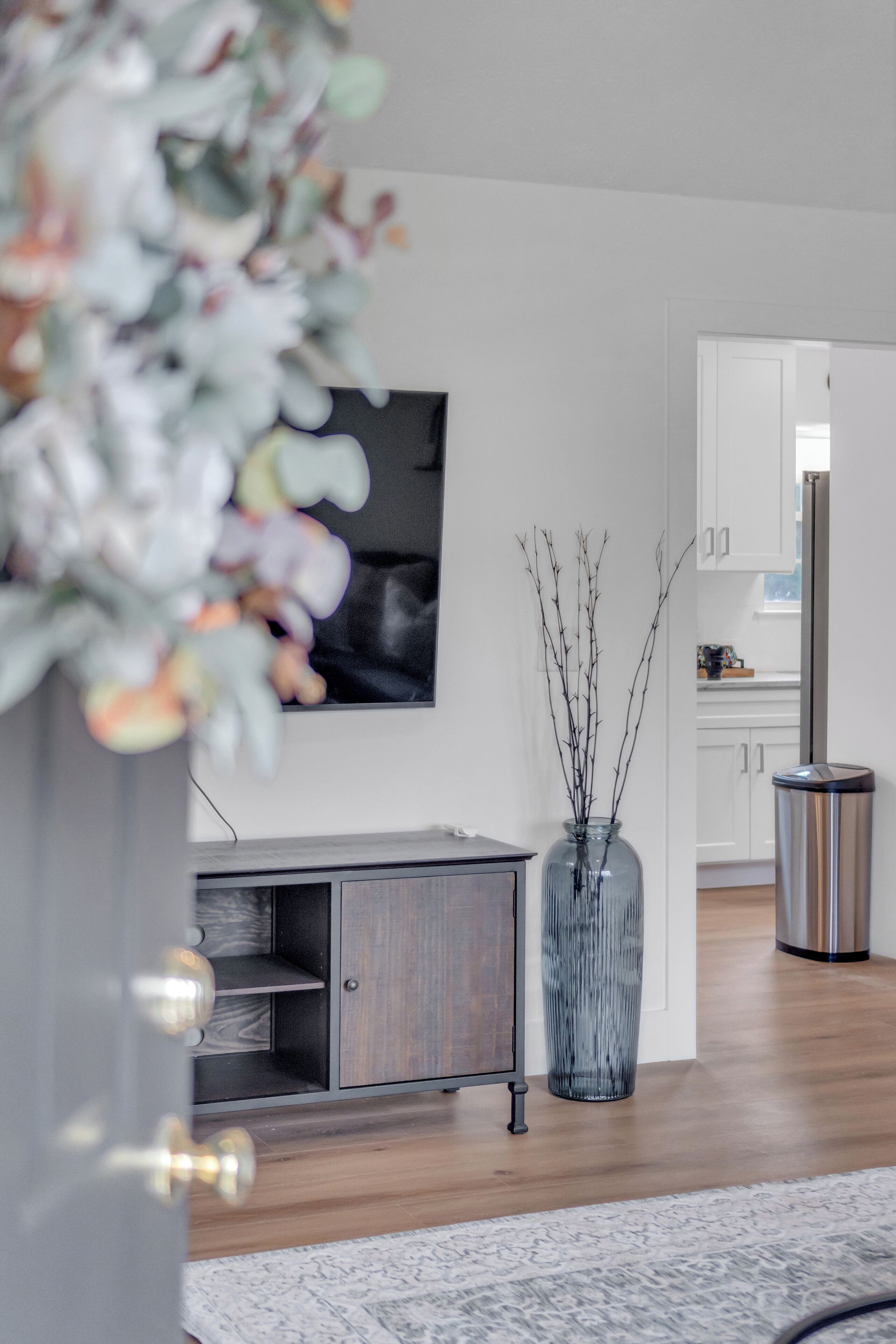 A modern living room with a wooden floor, a grey cabinet, a glass vase with branches, and a stainless steel trash bin.