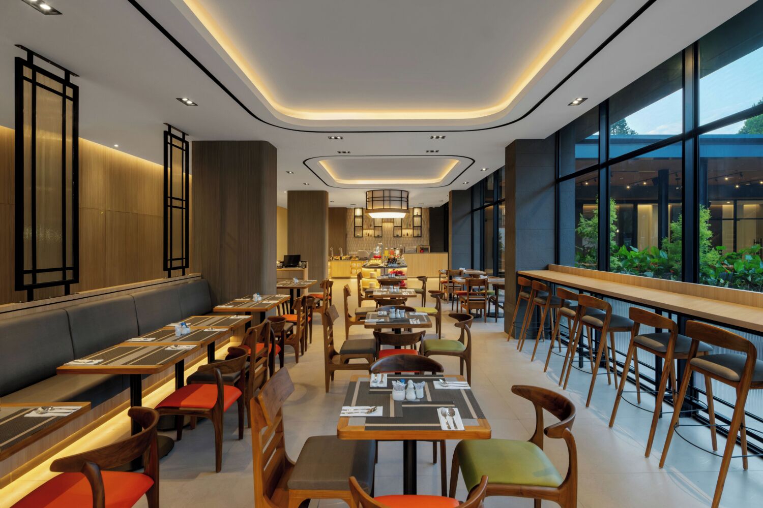A modern restaurant with a long counter, multiple tables, and chairs. There are large windows on one side and a kitchen area in the center.