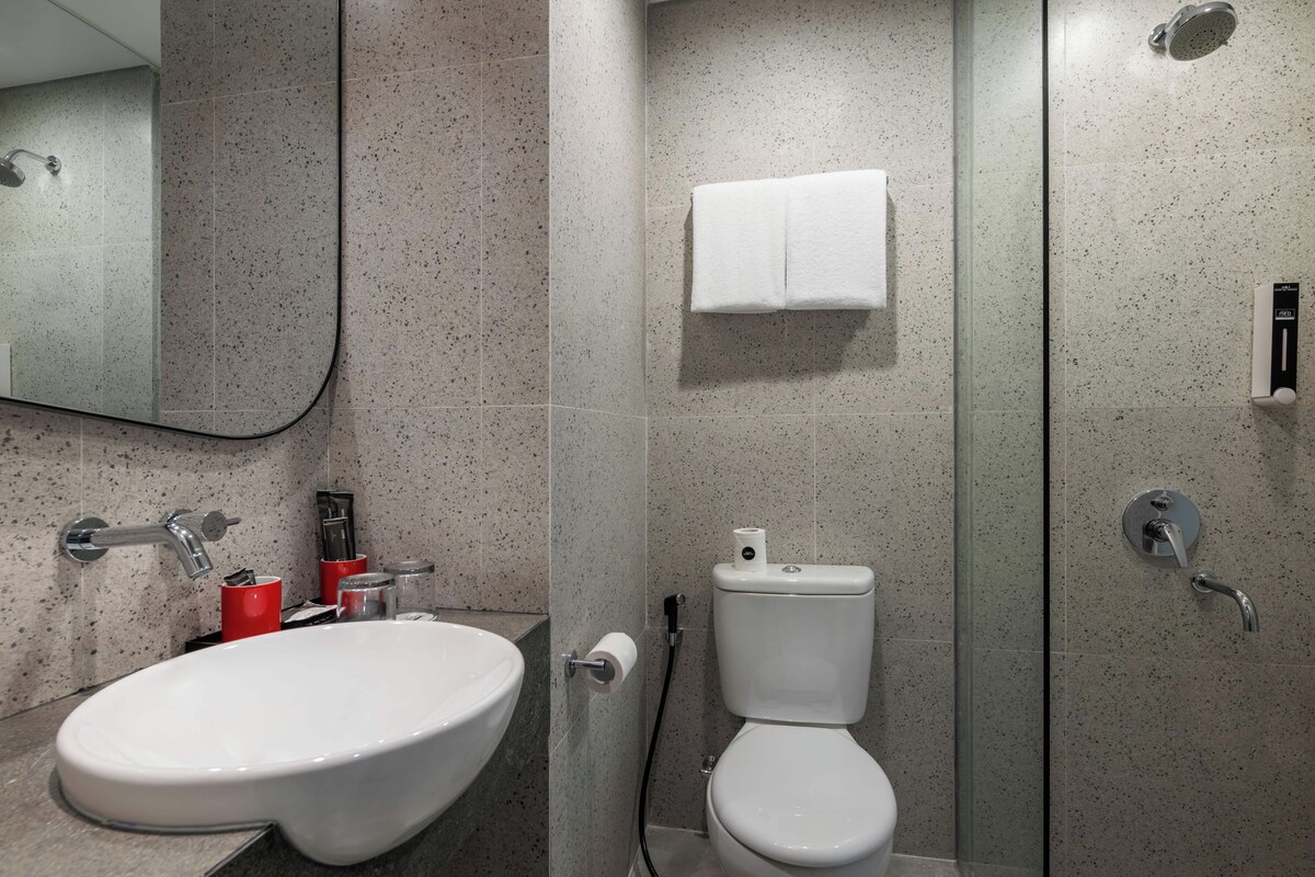 Ein Badezimmer mit weißem Waschbecken, WC und Dusche. Ein Handtuch hängt an der Wand, über dem Waschbecken befindet sich ein Spiegel.