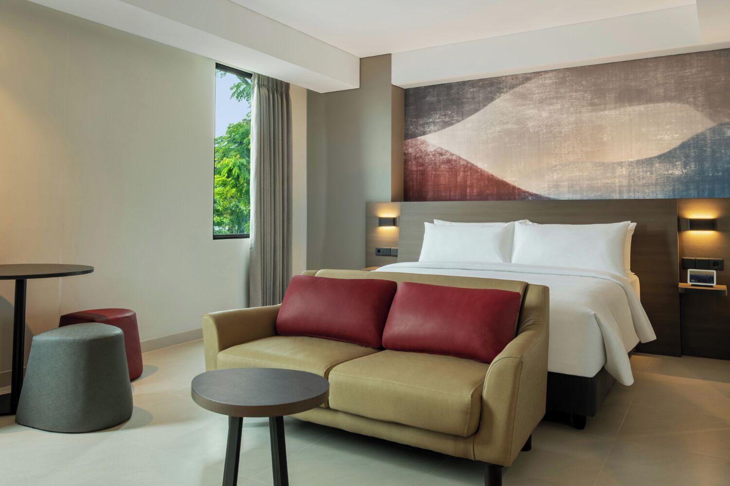 A modern hotel room with a bed, a sofa, a small table, and a painting on the wall.