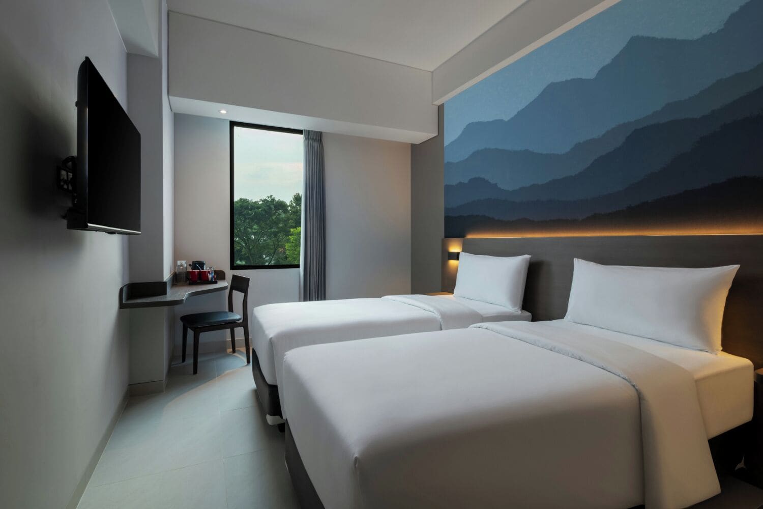 Habitación de hotel con dos camas, un escritorio, una silla, un televisor y un ventanal con vistas a árboles.