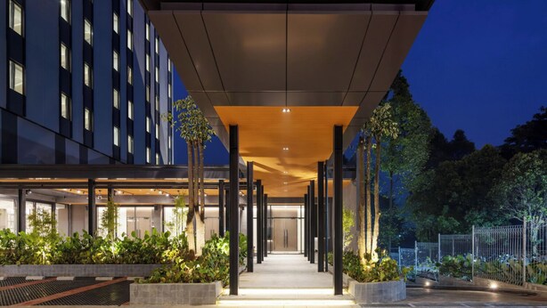 Une entrée d’hôtel moderne, dotée d’une allée couverte, de grandes fenêtres et d’un chemin bien éclairé menant au bâtiment.