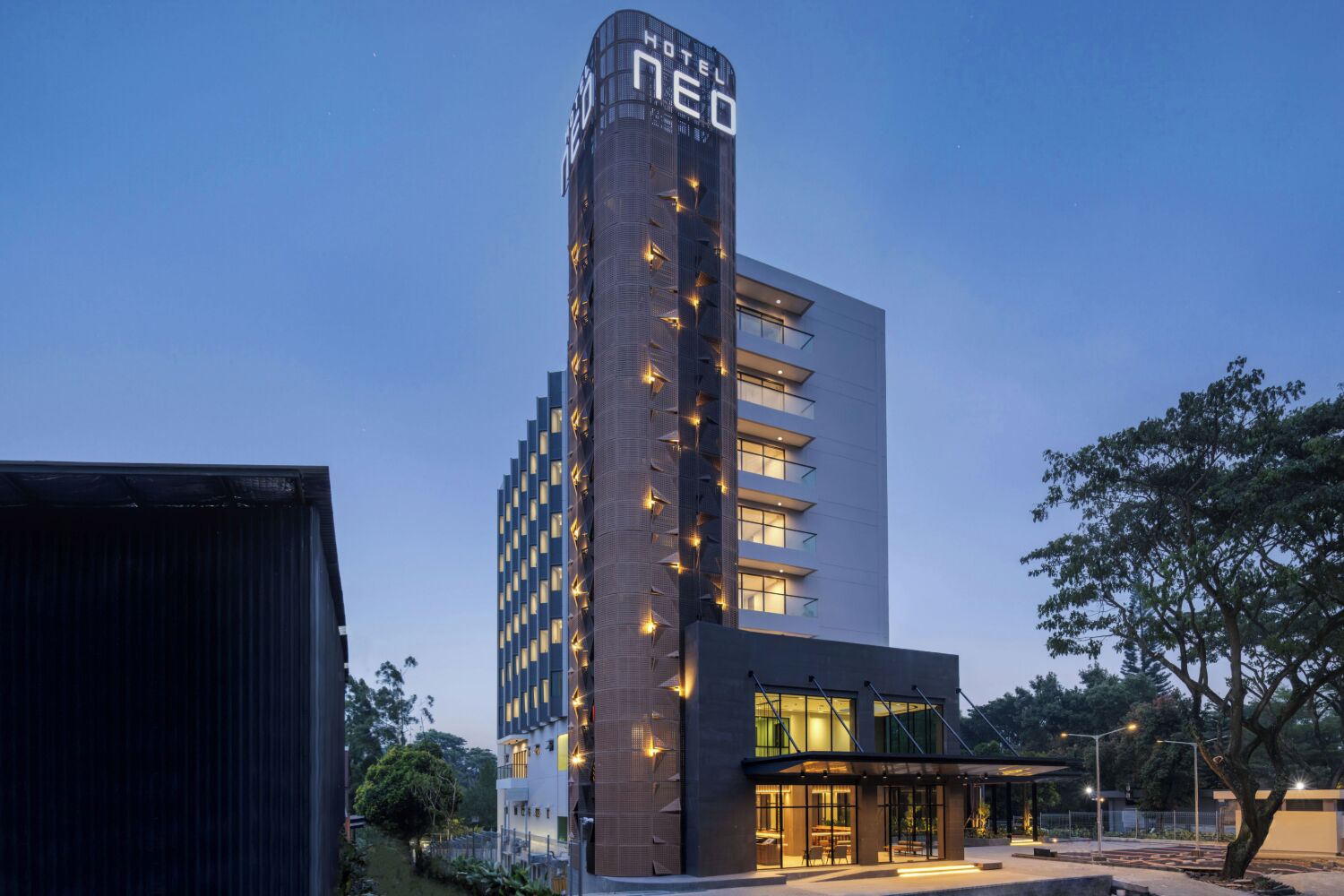 Un hôtel moderne doté d’une haute tour cylindrique sur laquelle on peut lire l’inscription « HÔTEL NEO », et d’une entrée au niveau du sol avec des portes en verre et un espace abrité.