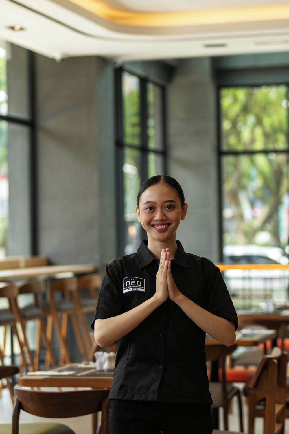 A person in a black uniform with a logo, standing in a modern indoor setting with wooden tables and chairs, large windows, and a view of greenery outside.