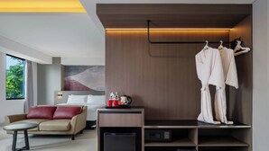 Une chambre d’hôtel moderne comprenant un lit, un canapé, une table basse, une armoire avec des vêtements suspendus et un porte-chaussures.