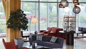 A modern lounge area with a grey sofa, red armchair, and round coffee table. A large wooden column with a plant, a metal sculpture, and a view of greenery outside. - Kinock Pet Hotel (Gyeongju)