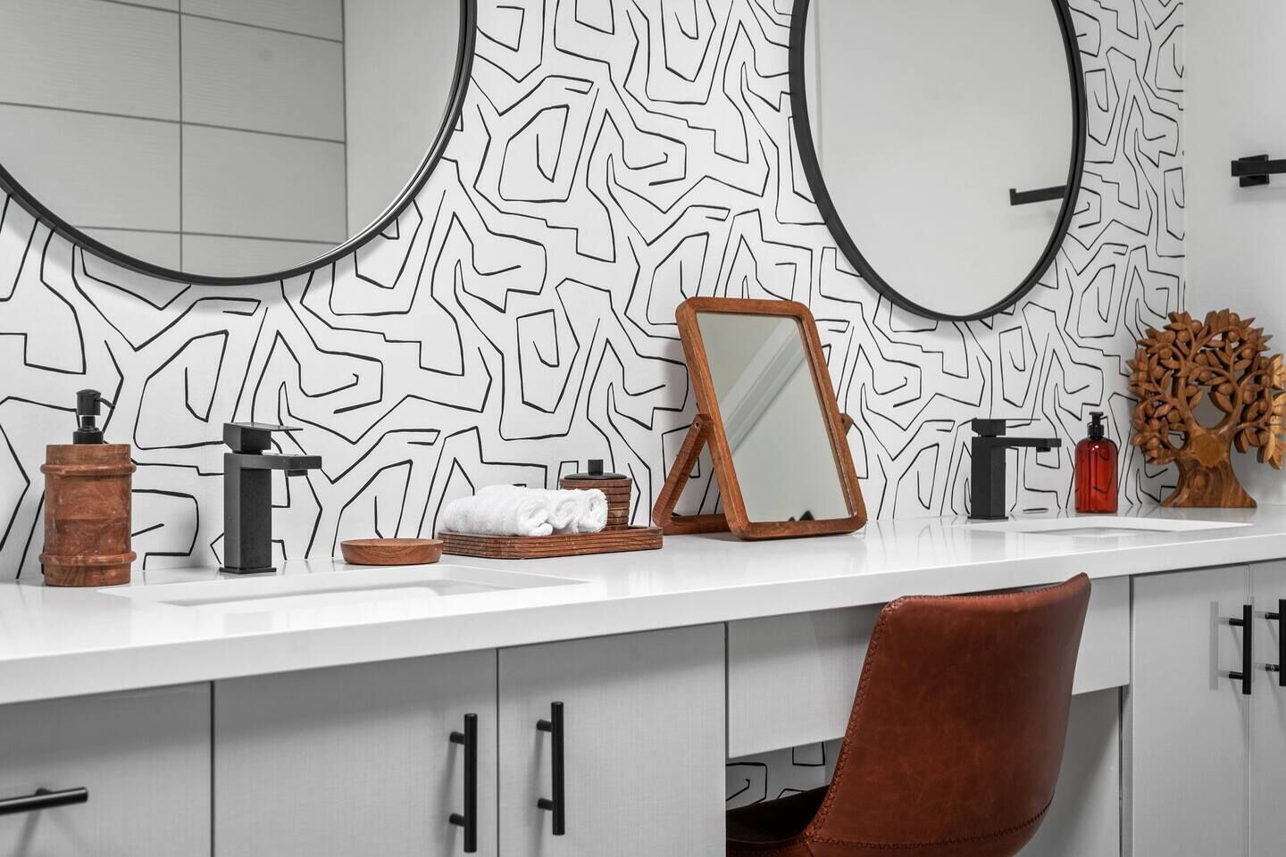 A bathroom with a white vanity, two mirrors, a wooden frame, a towel rack, a soap dispenser, and a decorative tree.