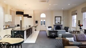 A modern kitchen with a marble island, a dining area with a round table, a living room with a sofa and fireplace, and large windows with blinds. - Penthouse Ozone in Queenscliff (Queenscliffe)