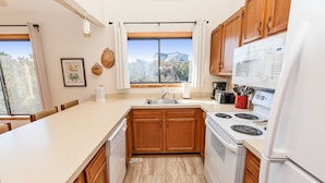 A modern kitchen with wooden cabinets, a white countertop, a built-in oven, and a window with a view of trees and a house. - Oceanside, Close to beach, Hot Tub, Dogs Welcome-CI2, Brass Ring (Duck)