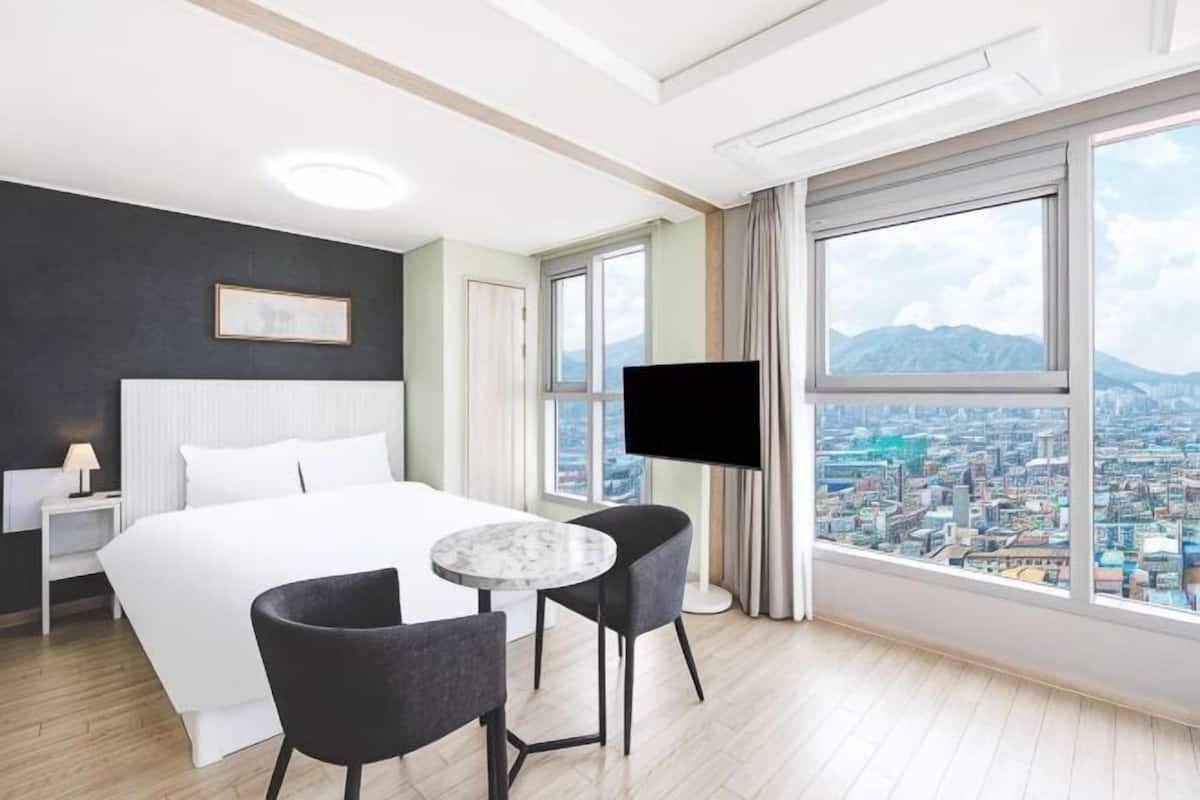 A hotel room with a bed, two chairs, a TV, and a large window with a city view.