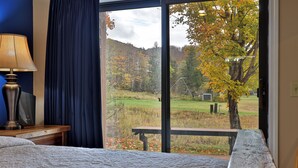 Ein Schlafzimmer mit großem Fenster, einem Bett, einem Nachttisch mit Lampe und Blick auf einen Park mit Bäumen und einem Picknicktisch.