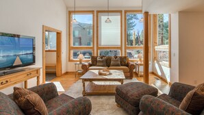 A spacious living room with a large TV, wooden furniture, and large windows offering a view of the outdoors. - Sparrow's Rest by Tahoe Getaways - Dog Friendly 3BR in Tahoe Donner w/ HOA (Truckee)