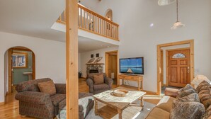 A spacious living room with a high ceiling, wooden flooring, and a cozy fireplace. There is a television on a wooden stand and a comfortable seating area with sofas and a coffee table. - Sparrow's Rest by Tahoe Getaways - Dog Friendly 3BR in Tahoe Donner w/ HOA (Truckee)