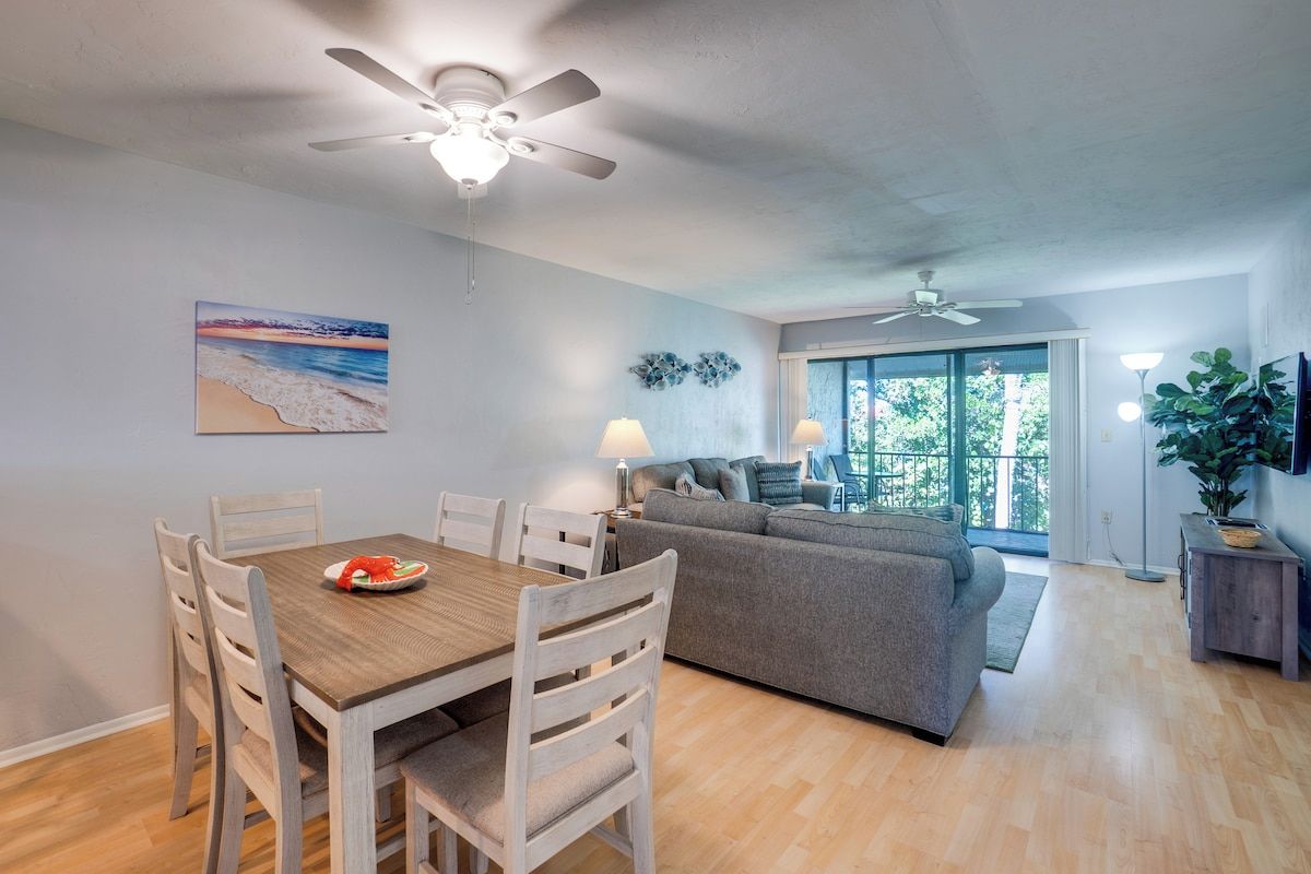 A dining area with a wooden table and chairs, a ceiling fan, and a painting of a beach scene.