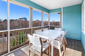 Um deck coberto ao ar livre com uma mesa e cadeiras brancas, guarda-corpos de madeira e vista para casas e árvores.