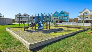 A playground with a slide and climbing structures, surrounded by a wooden fence and a grassy area.