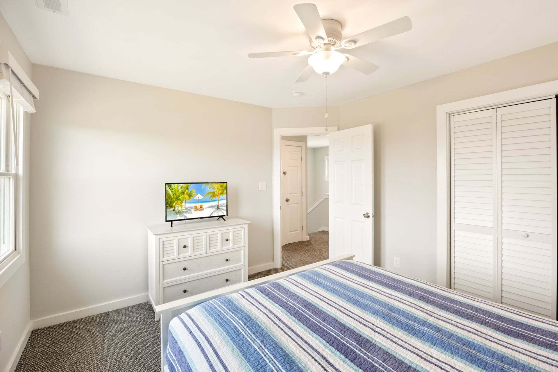 A bedroom with a bed, a television on a dresser, a ceiling fan, and a door leading to another room.