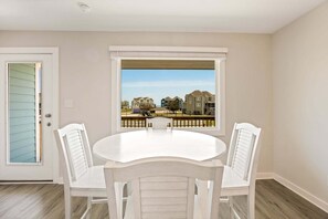 A dining area with a round table and four white chairs, a door with a view to a residential area, and a ceiling light fixture. - Pollock: Bright Buxton Townhouse w/ Ocean Views (Buxton)