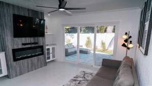 Un salon moderne avec un canapé gris, un téléviseur à écran plat fixé à un mur en bois, un ventilateur de plafond et une porte-fenêtre coulissante donnant sur une terrasse extérieure.