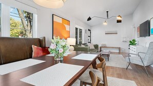 A modern living room with a wooden dining table, leather sofa, and a green armchair. There is a large window with a view of trees and a painting on the wall. - Boxwoods | Pool | Beautiful Interior (Fredericksburg)