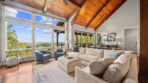 A spacious living room with a large beige sectional sofa, a coffee table, and a chandelier. There are multiple windows providing a view of the outdoors. - Resort Home–Scenic Views-Whispering Ridge Estate (Boone)