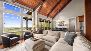 A spacious living room with a large sectional sofa, a coffee table, and a fireplace. There are large windows offering a view of the outdoors. - Resort Home–Scenic Views-Whispering Ridge Estate (Boone)