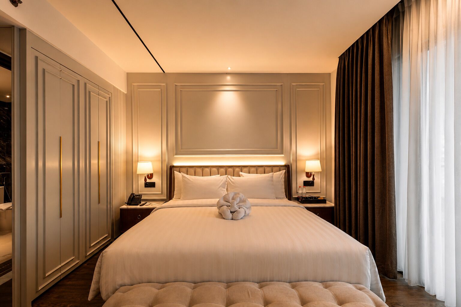 A hotel room with a large bed, two bedside tables with lamps, a headboard, and a large window with curtains.
