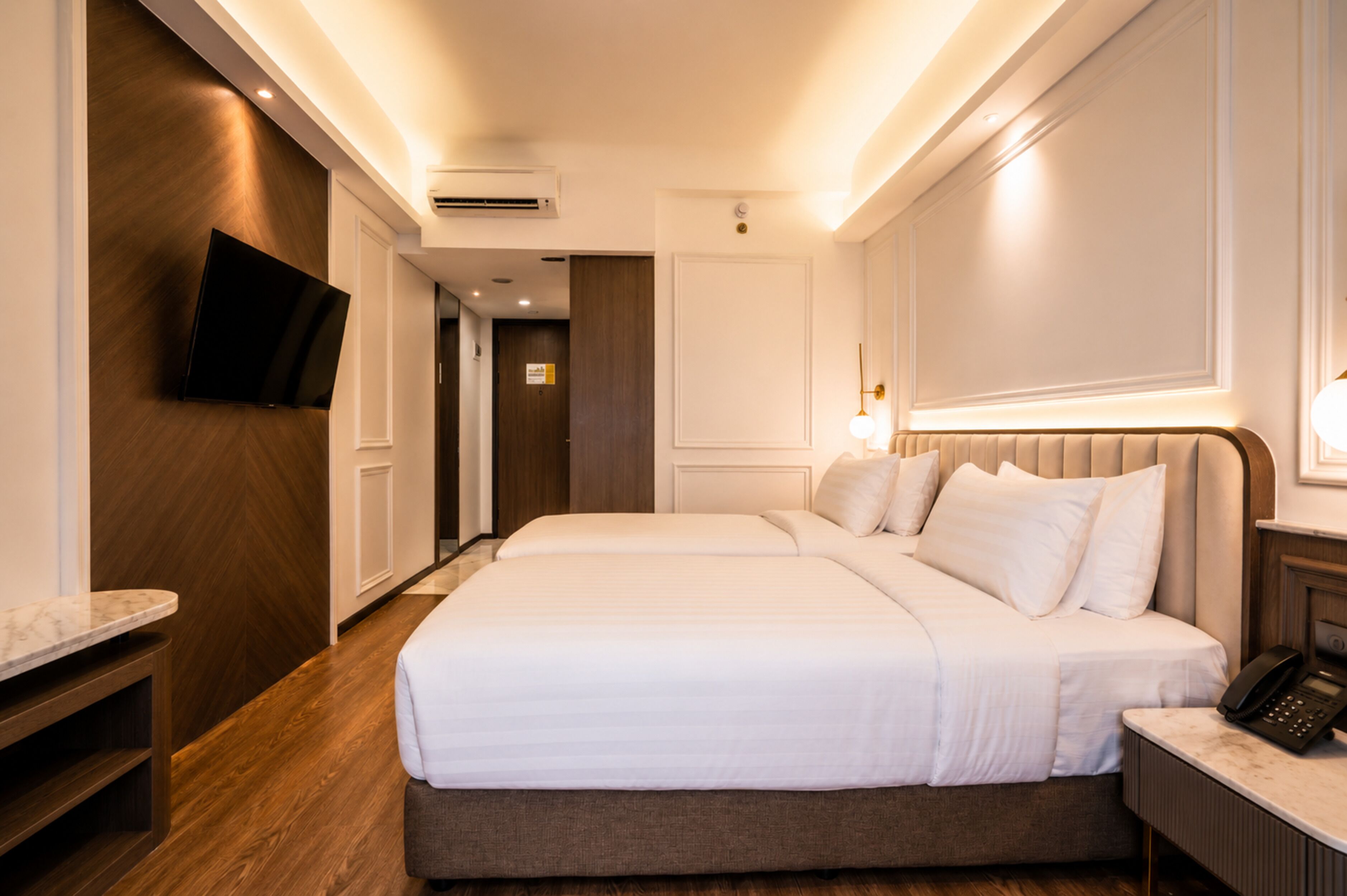 A hotel room with two beds, a flat-screen TV mounted on the wall, a bedside telephone, and a wooden floor.