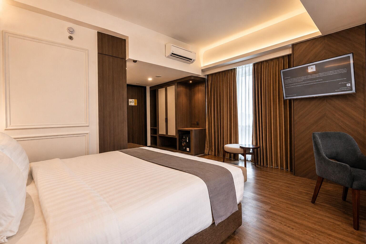 A hotel room with a bed, a desk, a chair, an air conditioning unit, and a mounted screen.