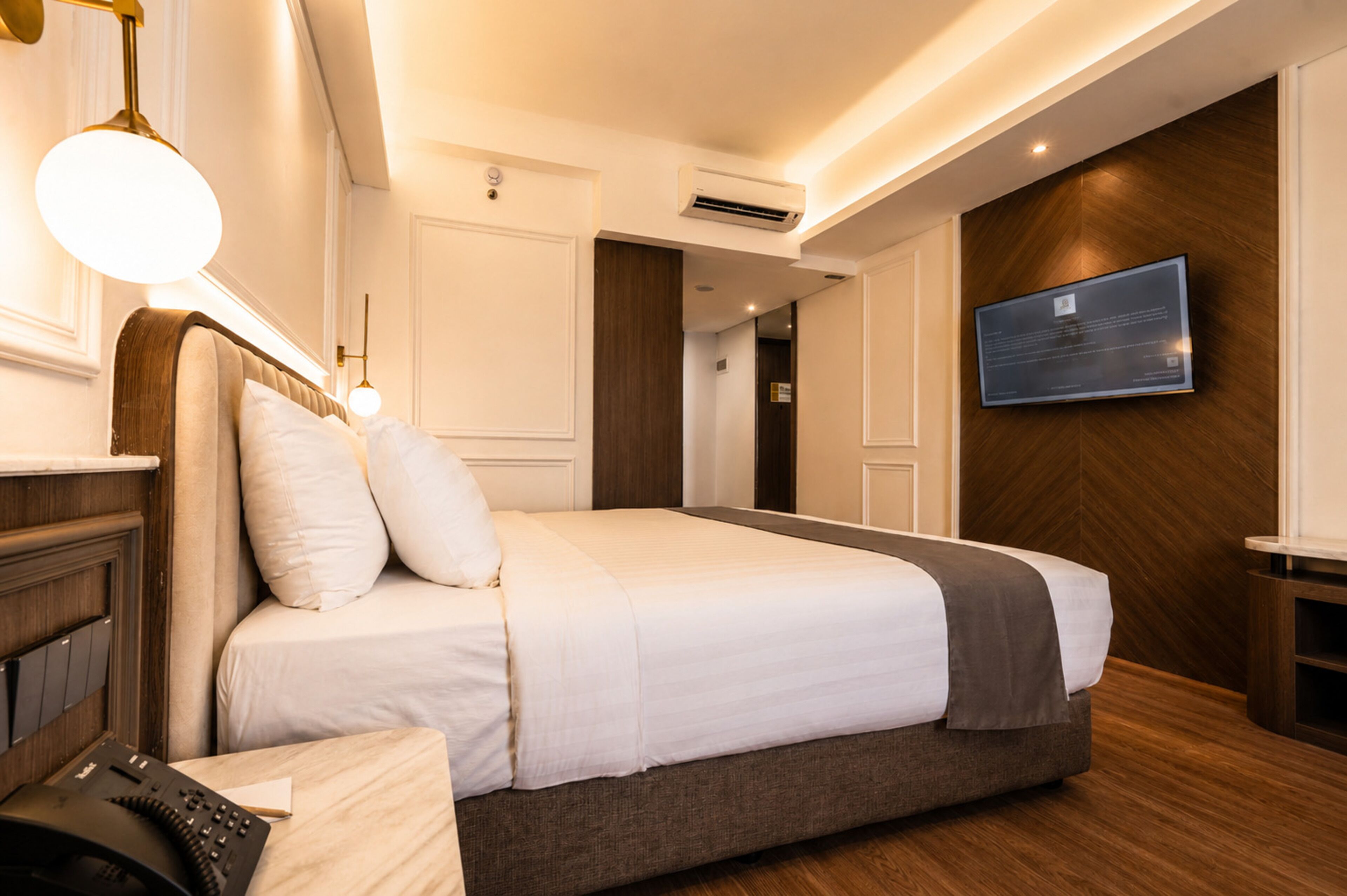 Ein Hotelzimmer mit einem großen Bett, einem flachbildfernseher an der Wand, einer Klimaanlage und einem Telefon auf dem Nachttisch.