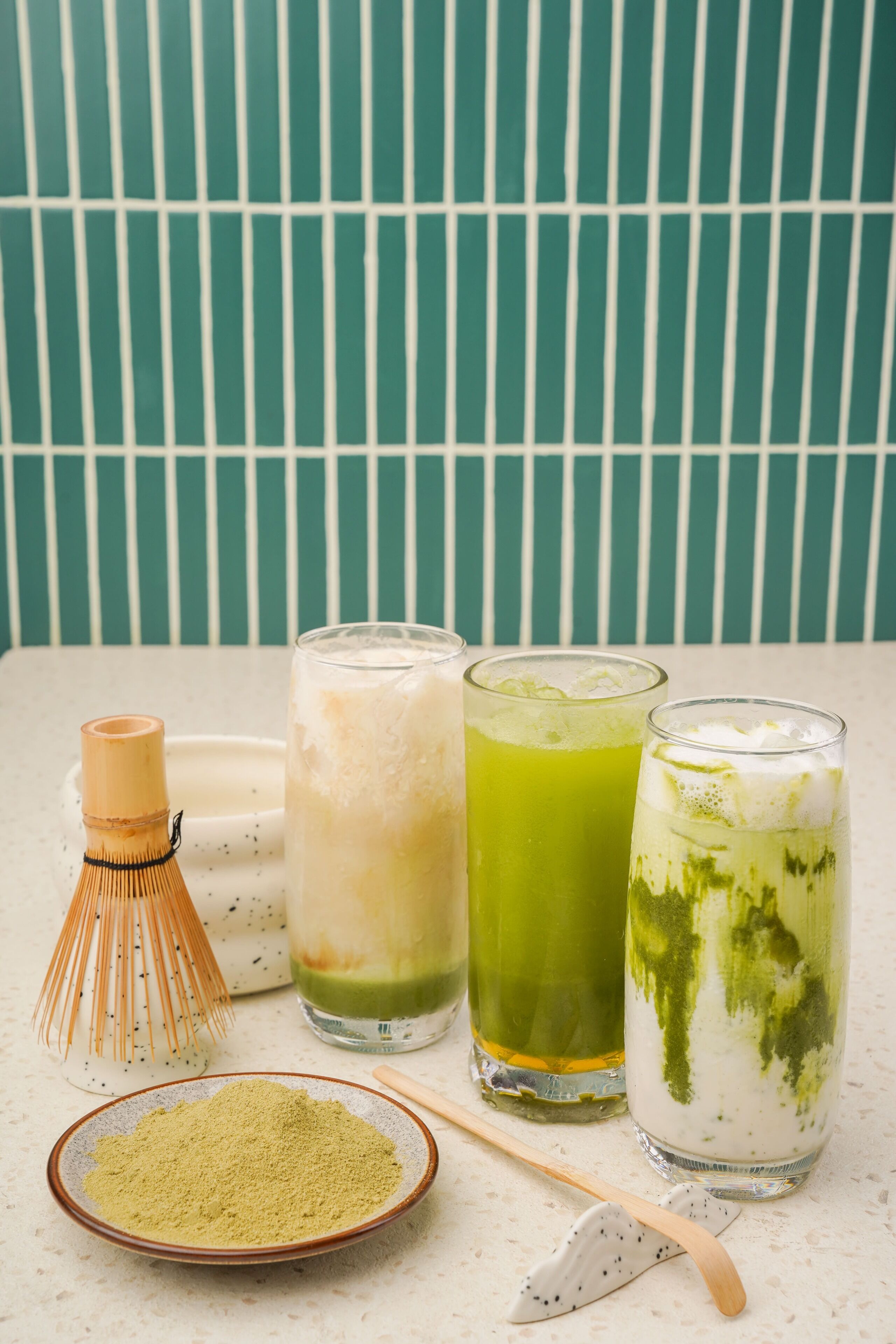 Trois verres de matcha sont posés sur une table, accompagnés d’un fouet en bambou et d’un bol de poudre de matcha.