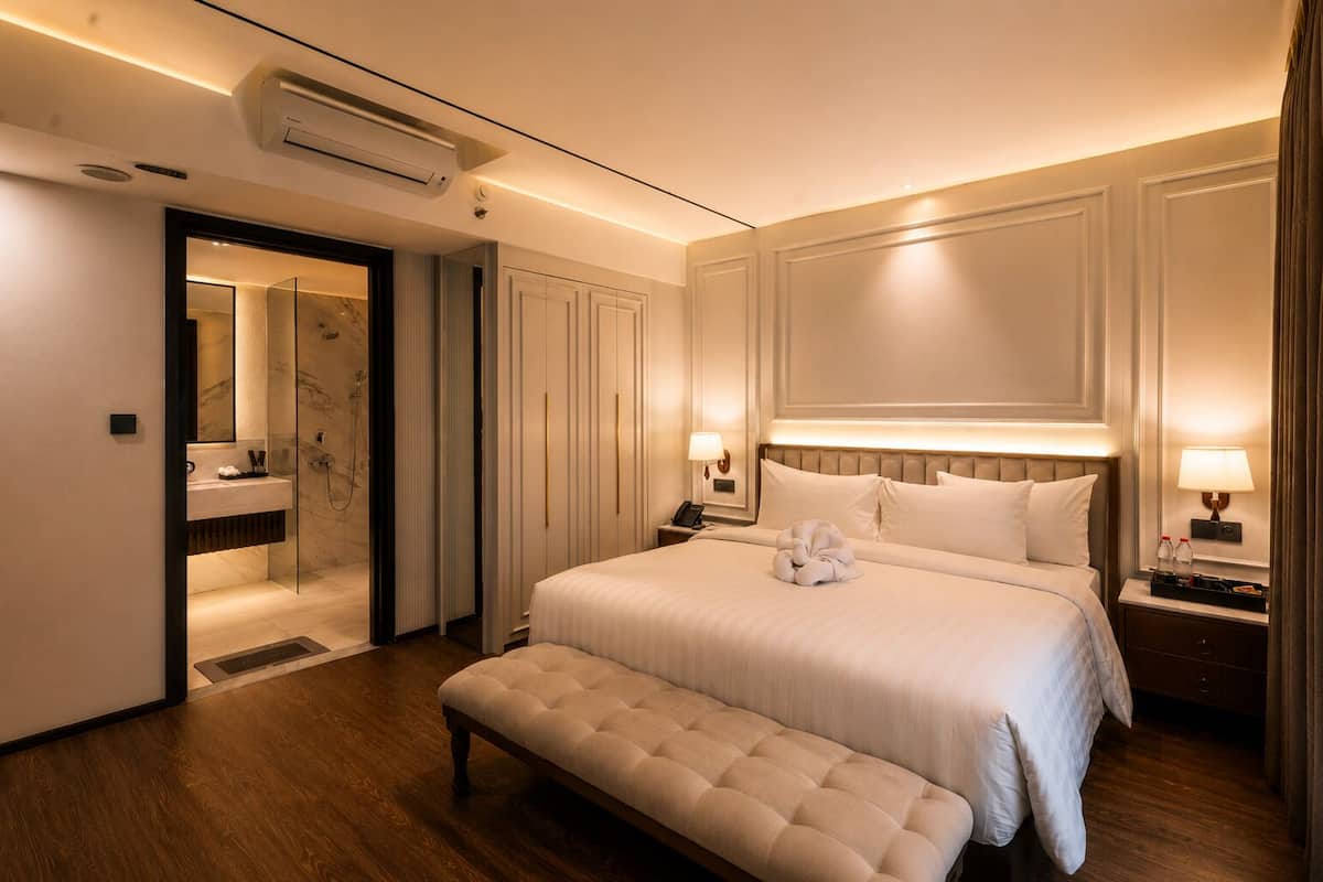 Quarto de hotel com uma cama grande, mesas de cabeceira, um banco, um espelho, chuveiro e um guarda-roupa.