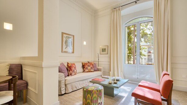 Ein helles Wohnzimmer mit einer Couch, einem Hocker, einem Sessel, einem Couchtisch und einem großen Fenster mit Vorhängen.