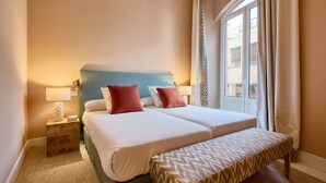 Ein Doppelbett mit zwei Kissen, ein Nachttisch mit Lampe, eine Bank und ein Fenster mit Vorhängen.