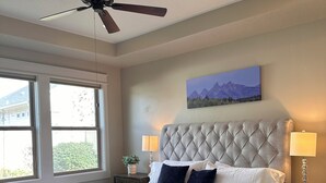 Une chambre à coucher avec une tête de lit capitonnée, un grand lit recouvert de draps blancs, deux lampes de chevet, un ventilateur de plafond et une fenêtre donnant sur un paysage verdoyant.