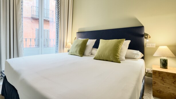 A hotel room with a large bed, a nightstand with a lamp, and a view of a building through a window. - Adriano 1.4 by Home at Homes (Sevilla)