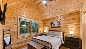 Ein Schlafzimmer einer Holzhütte mit Bett, Nachttischen, einem Beistelltisch, einer Bank, einem Deckenventilator und einem Fenster mit Blick auf den Wald.