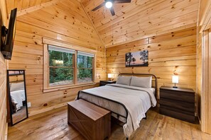 Quarto de cabana de madeira com uma cama, mesas de cabeceira, um criado-mudo, um banco, um ventilador de teto e uma janela com vista para a floresta.