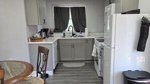Une cuisine compacte avec un coin repas, des meubles blancs, une poubelle noire et un réfrigérateur.