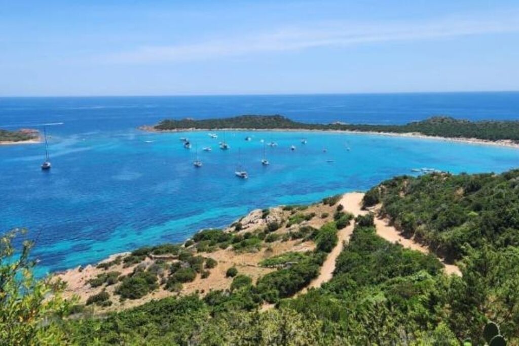 A coastal landscape with clear blue waters, boats, and a hilly terrain with vegetation.