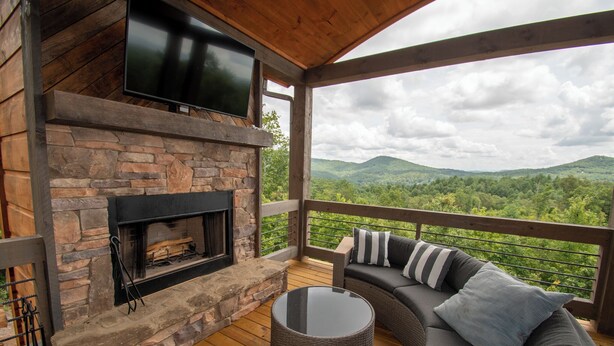 A covered outdoor area with a stone fireplace, a flat-screen TV, a cushioned seating area, and a view of green hills.