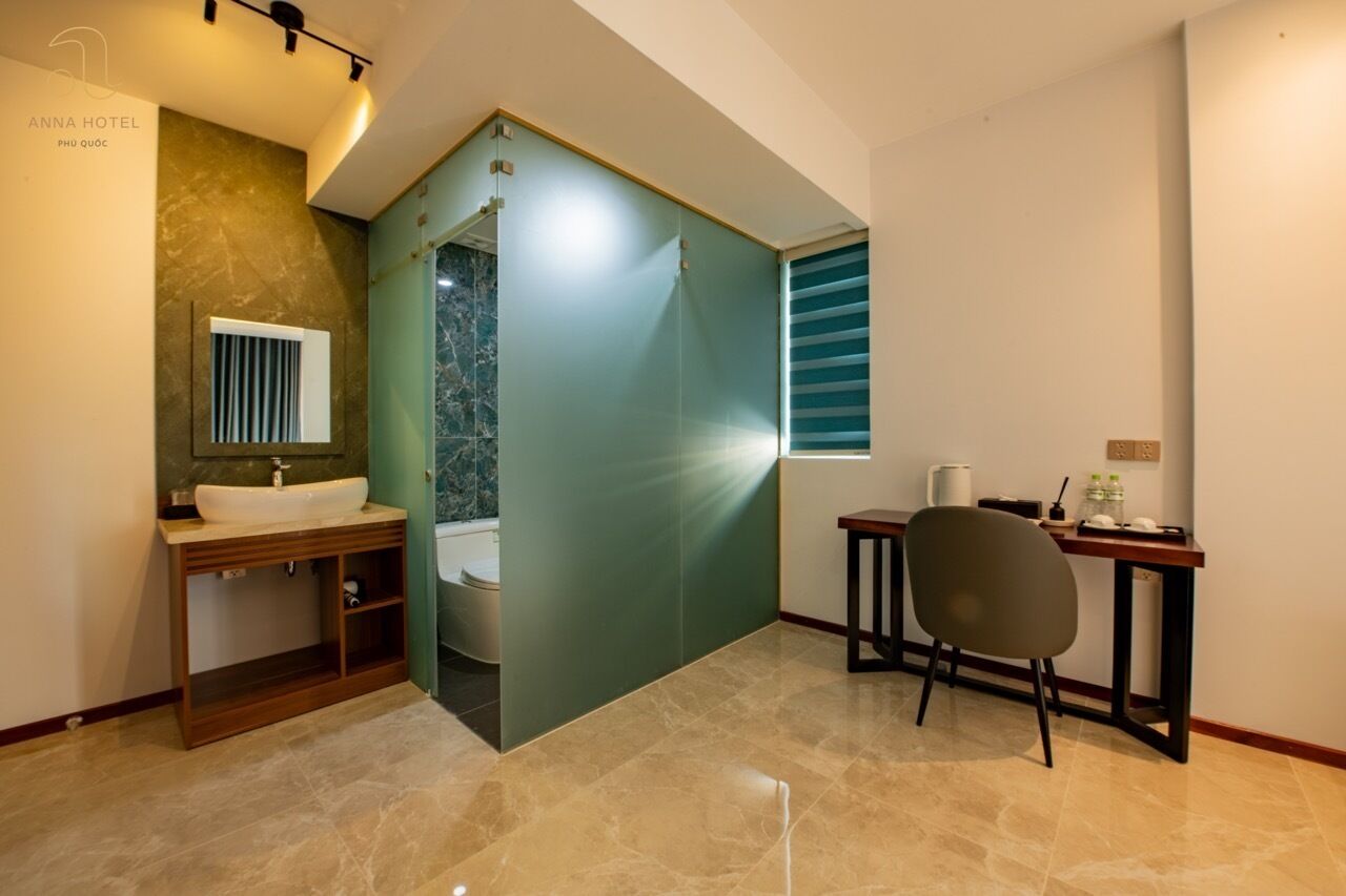 Un baño pequeño con tocador, lavabo e inodoro. Hay una mesita con una silla y una ventana con persianas.