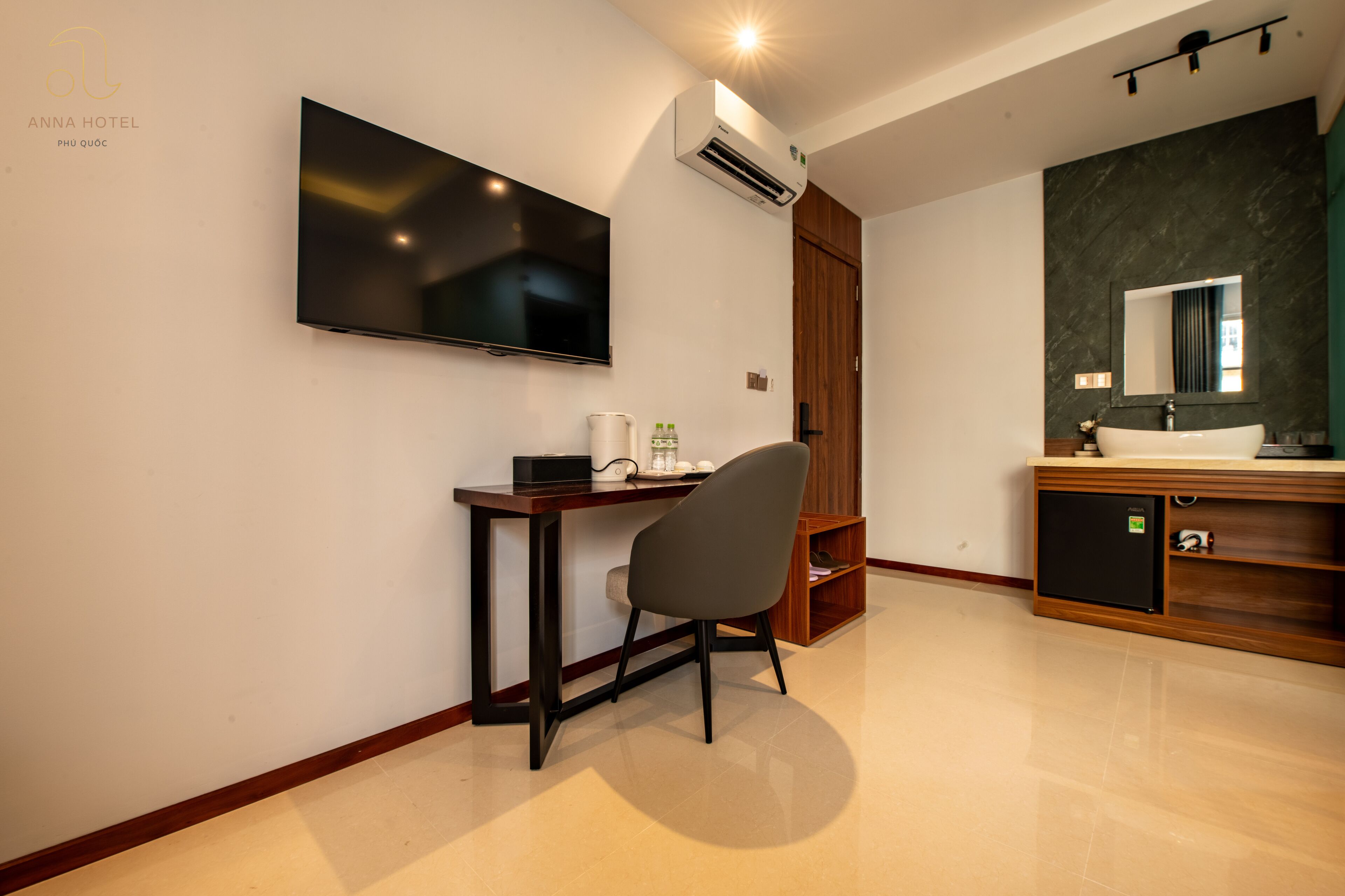 A modern hotel room with a flat-screen TV, a desk with a chair, a wooden wardrobe, a built-in sink, and a mirror.