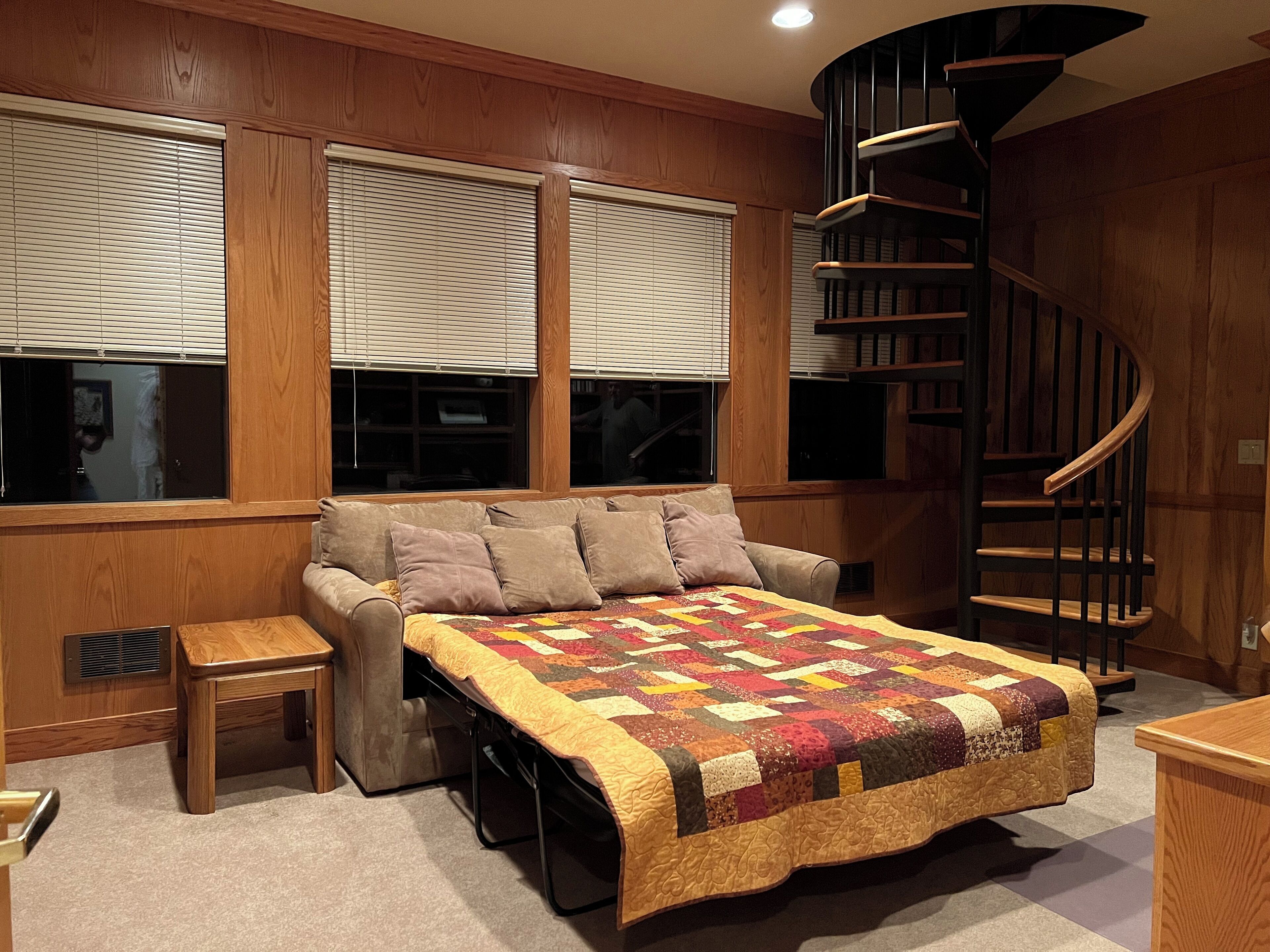 A bedroom with a bed, a small table, a chair, a window with blinds, and a spiral staircase.