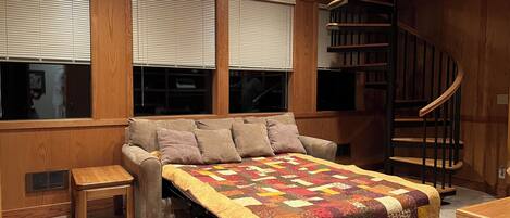 A bedroom with a bed, a small table, a chair, a window with blinds, and a spiral staircase.