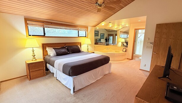 Un dormitorio con cama, lámparas de noche, techo de madera, un ventanal grande y un baño visible a través de una puerta abierta.