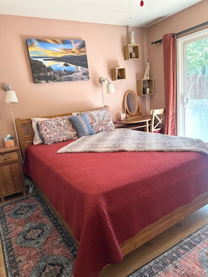 A bedroom with a wooden bed, a red bedspread, a patterned pillow, a bedside table with a lamp, a mirror, and a view of the outdoors through a window. - Pinter Pines | Hot Tub | BBQ | Mini Golf (South Lake Tahoe)