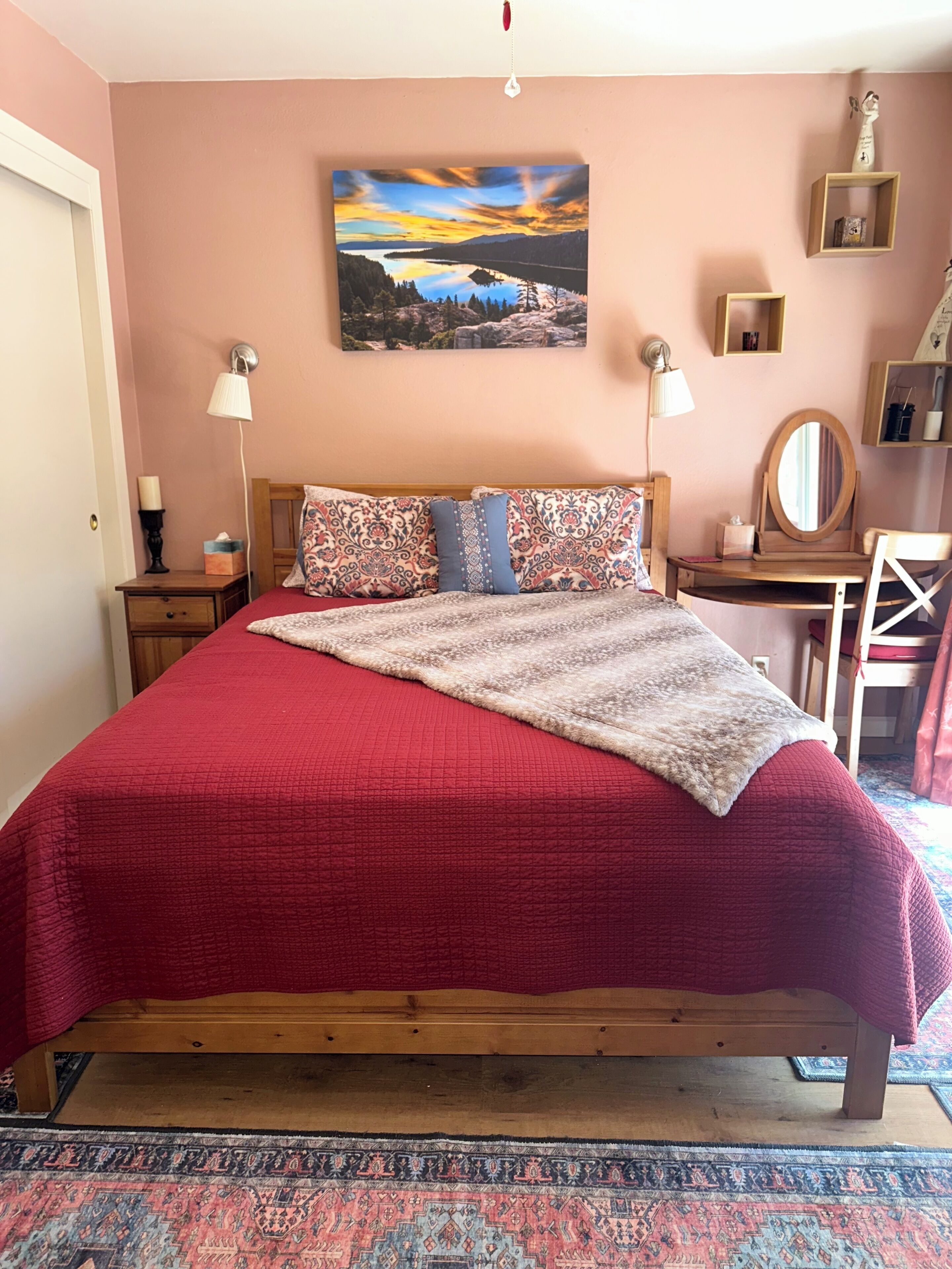 Ein Schlafzimmer mit einem hölzernen Bett, einer roten Bettdecke, einem gemusterten Kopfkissen, einer Nachttischlampe, einem Nachttisch aus Holz, einem Wandbild und einem kleinen Schreibtisch mit Spiegel.